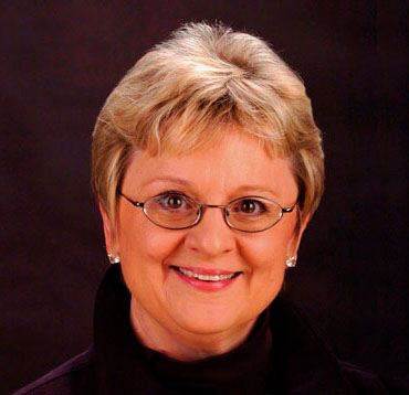 a white woman with short blonde hair and glasses wearing a black shirt in front of a dark background