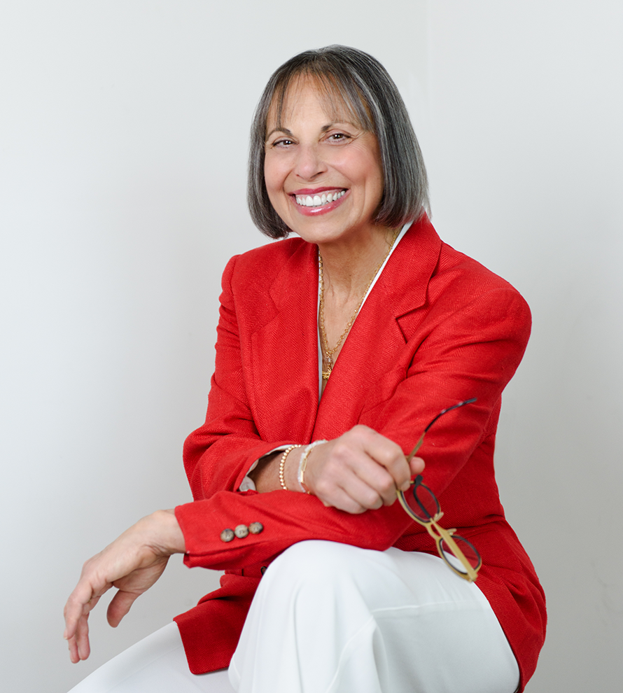 Judy Baker is a white woman with salt and pepper shoulder length hair. She's wearing a red blazer, white pants, and is smiling widely.