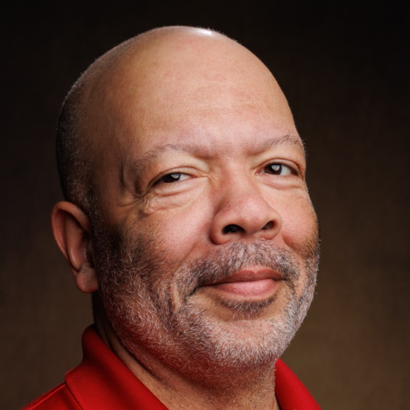 a black man with a bald head smiling gently into the camera wearing a red shirt.